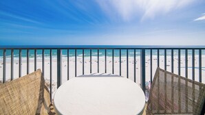 Een balkon met rieten stoelen en een ronde tafel, met uitzicht op een strand met mensen en een heldere lucht.