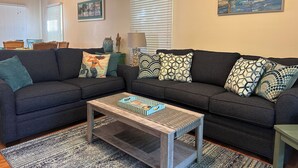 A living room with a dark grey sofa, patterned cushions, a wooden coffee table, and a framed painting of boats on water. -  Casa Capri Beautiful Tropical with 3 King Suites - 2 miles to beach (Key West)