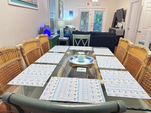Un coin repas avec une table à plateau de verre, des sets de table à motifs, des chaises en osier et, en arrière-plan, une vue sur le salon.
