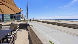 A coastal walkway with a beach in the background, a palm tree, and a building with outdoor seating. - Indoor/Outdoor living on the ocean front. (San Diego)