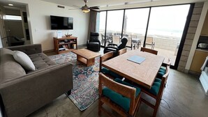 A modern living room with a sofa, coffee table, and dining table with chairs. There is a TV mounted on the wall and a large window overlooking the beach. - Indoor/Outdoor living on the ocean front. (San Diego)