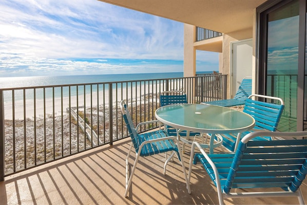 Un balcon avec une table en verre et des chaises bleues, donnant sur une plage et l’océan.
