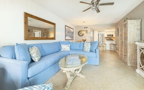 A living room with a blue sectional sofa, a glass coffee table, a ceiling fan, and a kitchen in the background.