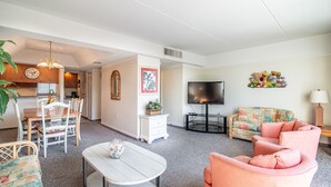 A living room with a sofa, a coffee table, and a TV. There is a dining area with a table and chairs. The room has a white ceiling and walls.