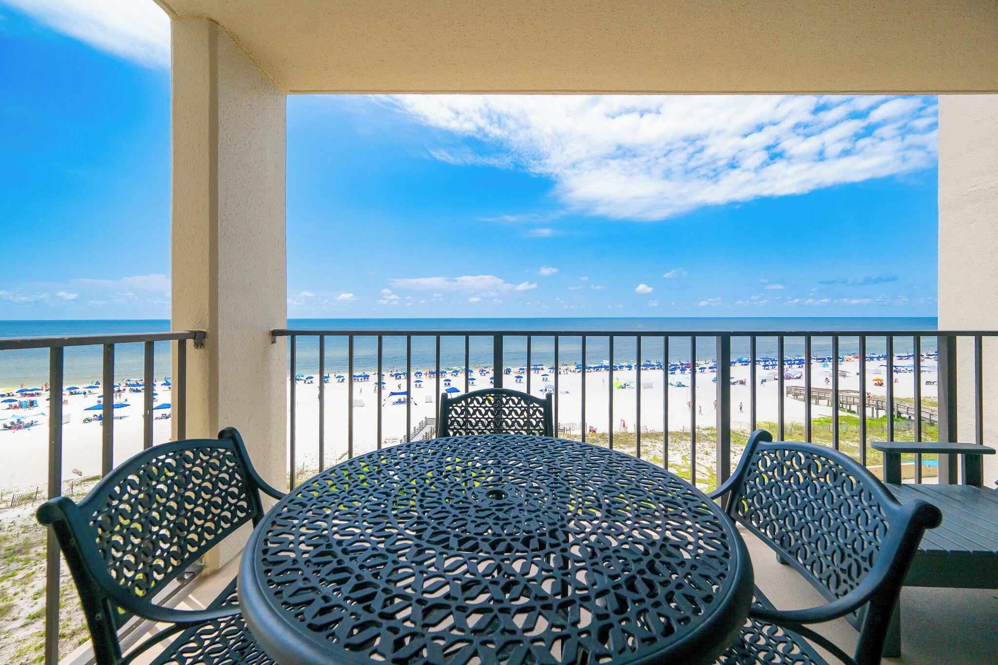 A balcony with a table and chairs, overlooking a beach with umbrellas and people.