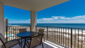 A balcony with a table and chairs, overlooking a beach with a clear view of the ocean and a distant building.