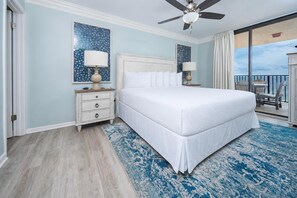 Quarto de hotel com uma cama grande, um ventilador de teto, mesa de cabeceira com abajur e vista para o mar.