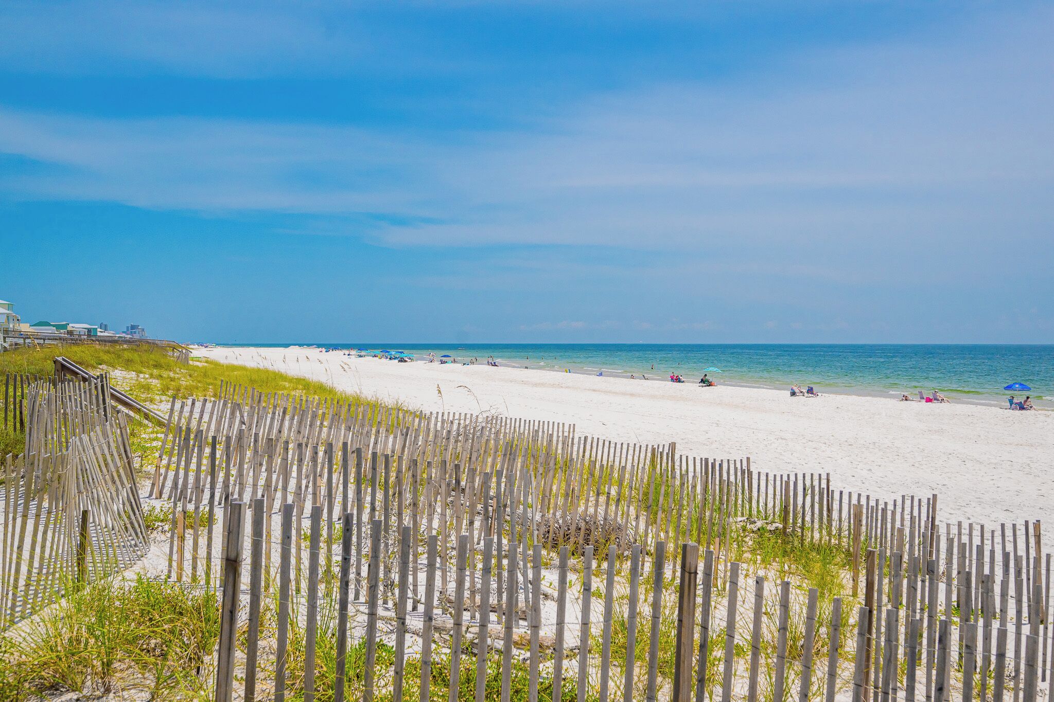 A beach with a wooden fence in the foreground, a sandy shore, and a clear blue sky.