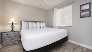 Ein Schlafzimmer mit Bett, Nachttisch, Lampe, Fenster mit Jalousien und zwei gerahmten Bildern an der Wand.