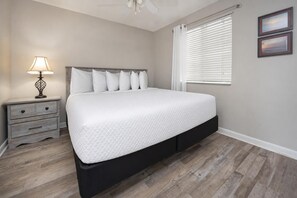 Un dormitorio con cama, mesita de noche, lámpara, ventana con persianas y dos cuadros en la pared.