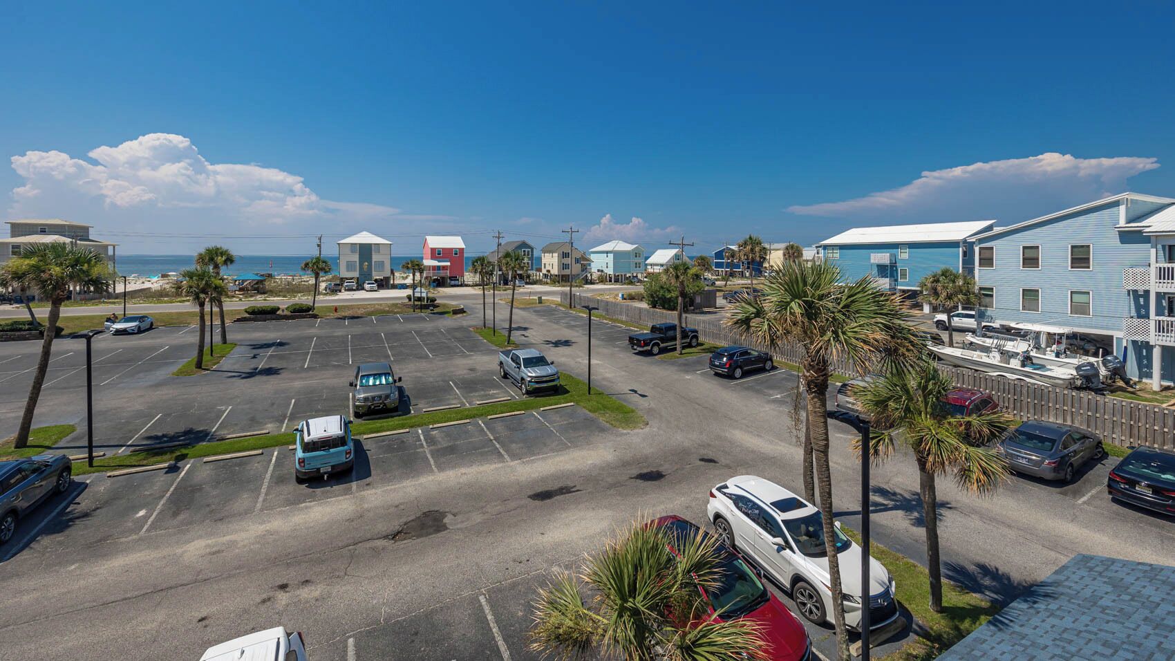 A parking lot with several cars, palm trees, and buildings with a beach view.