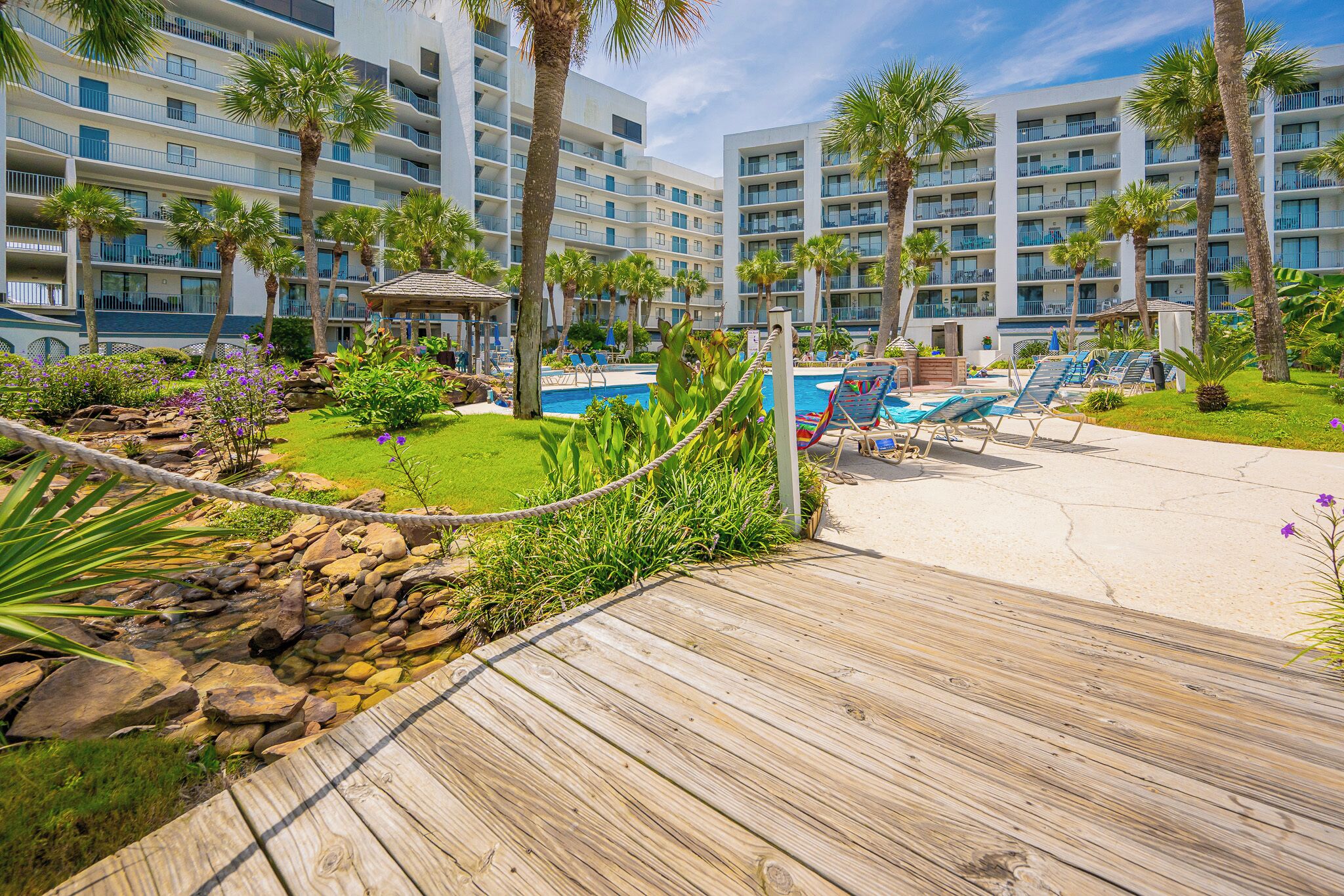 Un resort con un paseo de madera que conduce a una zona de piscina, rodeado de palmeras y un cielo despejado.