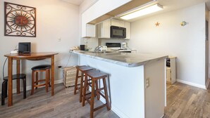Eine kompakte Küche mit einer zentralen Kochinsel, einer Mikrowelle und einem Spülbecken. Ein Holztisch mit zwei Hocker und einem dekorativen Wandbild sind ebenfalls vorhanden.
