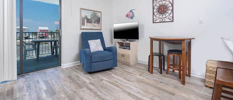 A living room with a blue armchair, a TV, a small wooden table, and a view of a balcony with outdoor furniture.