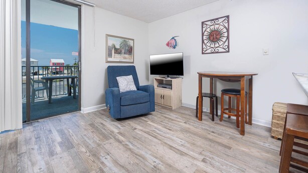 Ein Wohnzimmer mit blauem Sessel, Fernseher, kleinem Holztisch und Blick auf einen Balkon mit Gartenmöbeln.