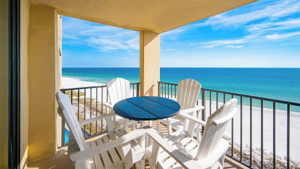 Een balkon met een ronde tafel en witte stoelen, met uitzicht op een strand met helderblauw water en een zandstrand.