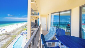 A balcony with a view of the beach, a pool, and a dining area with outdoor seating.