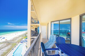 A balcony with a view of the beach, a pool, and a dining area with outdoor seating.