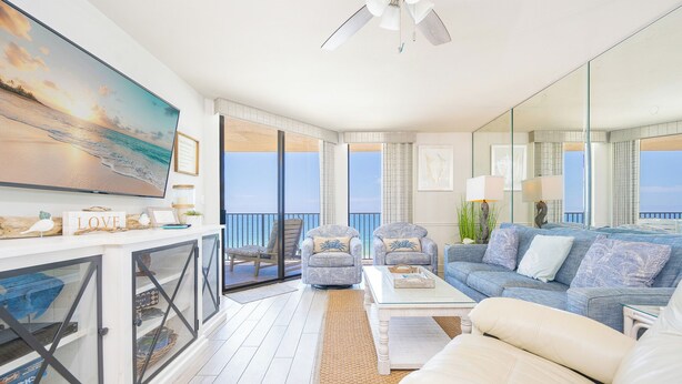 A spacious living area with a large TV, comfortable seating, a ceiling fan, and a balcony with a view.