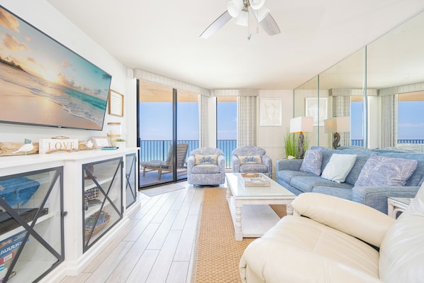 A spacious living area with a large TV, comfortable seating, a ceiling fan, and a balcony with a view.