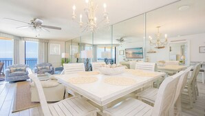 A spacious dining area with a large white table, white chairs, and a chandelier. There are ceiling fans and a large mirror reflecting the room.