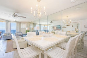 A spacious dining area with a large white table, white chairs, and a chandelier. There are ceiling fans and a large mirror reflecting the room.