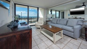 A spacious living room with a large window overlooking a beach, a grey sofa, a wooden coffee table, and a desk with a fish sculpture.