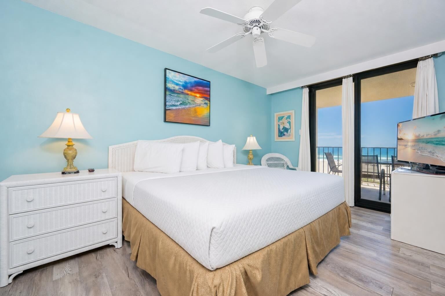 A hotel room with a bed, a nightstand, a lamp, a ceiling fan, a painting, a chair, a TV, and a balcony with a view.