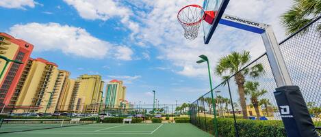 An outdoor basketball court with a net labeled "DOMINATOR," surrounded by a chain-link fence, palm trees, and buildings in the background.