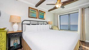 A bedroom with a bed, two wall-mounted paintings, a ceiling fan, a nightstand, a lamp, a window with a view of the ocean, and a wooden floor.