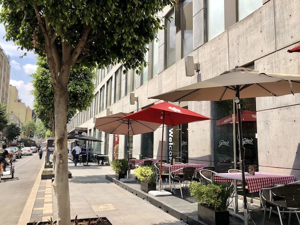 Un café en plein air avec des tables et des chaises, un arbre et un bâtiment doté de grandes fenêtres.