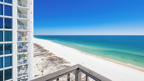 Ein Strandhotel mit Balkonen mit Blick auf einen Sandstrand und klares, blaues Wasser.