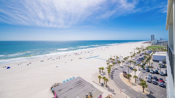 Blick auf einen Strand mit Parkplatz, Palmen und einem Gebäude im Hintergrund.