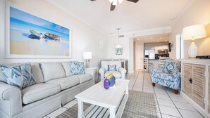 Un salon avec un canapé, une table basse et des fauteuils à motifs. On peut voir un tableau au mur, un ventilateur de plafond et, au fond, un coin cuisine.