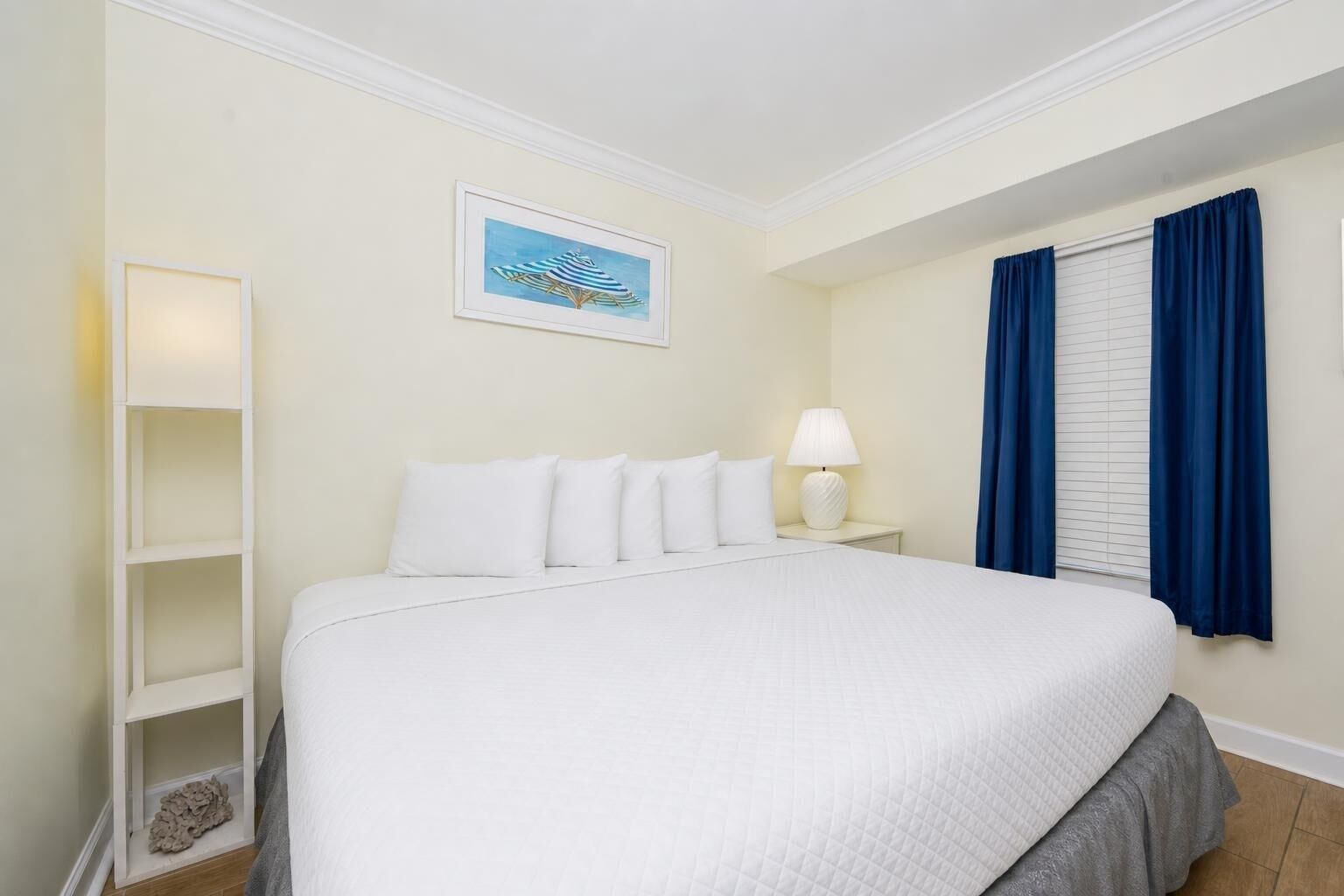 A hotel room with a large bed, a nightstand with a lamp, a framed picture on the wall, and a window with blue curtains.
