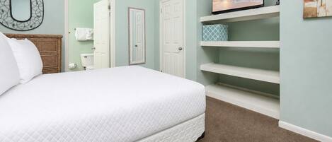 Un dormitorio con cama, televisor en un estante y baño con inodoro y perchero para toallas.