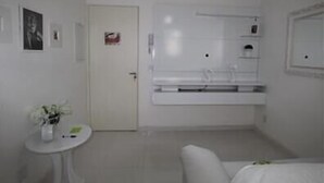 Baño pequeño y blanco de un hotel, con lavamanos blanco, espejo y una mesita redonda con un florero.