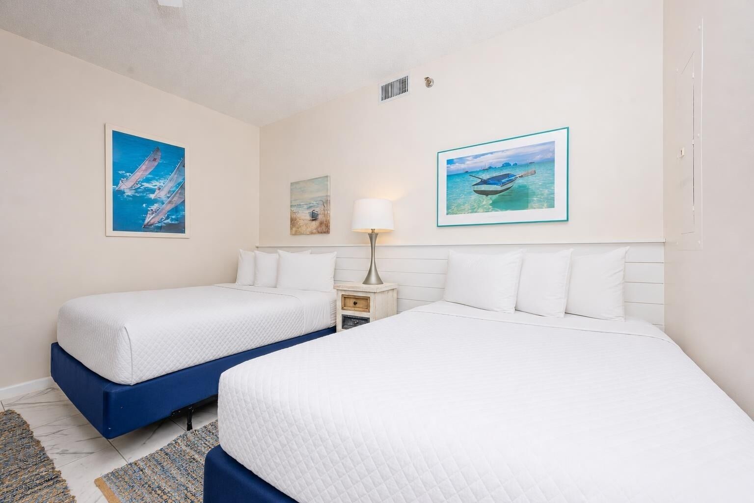 A hotel room with two beds, a nightstand, a lamp, and two framed pictures on the wall.