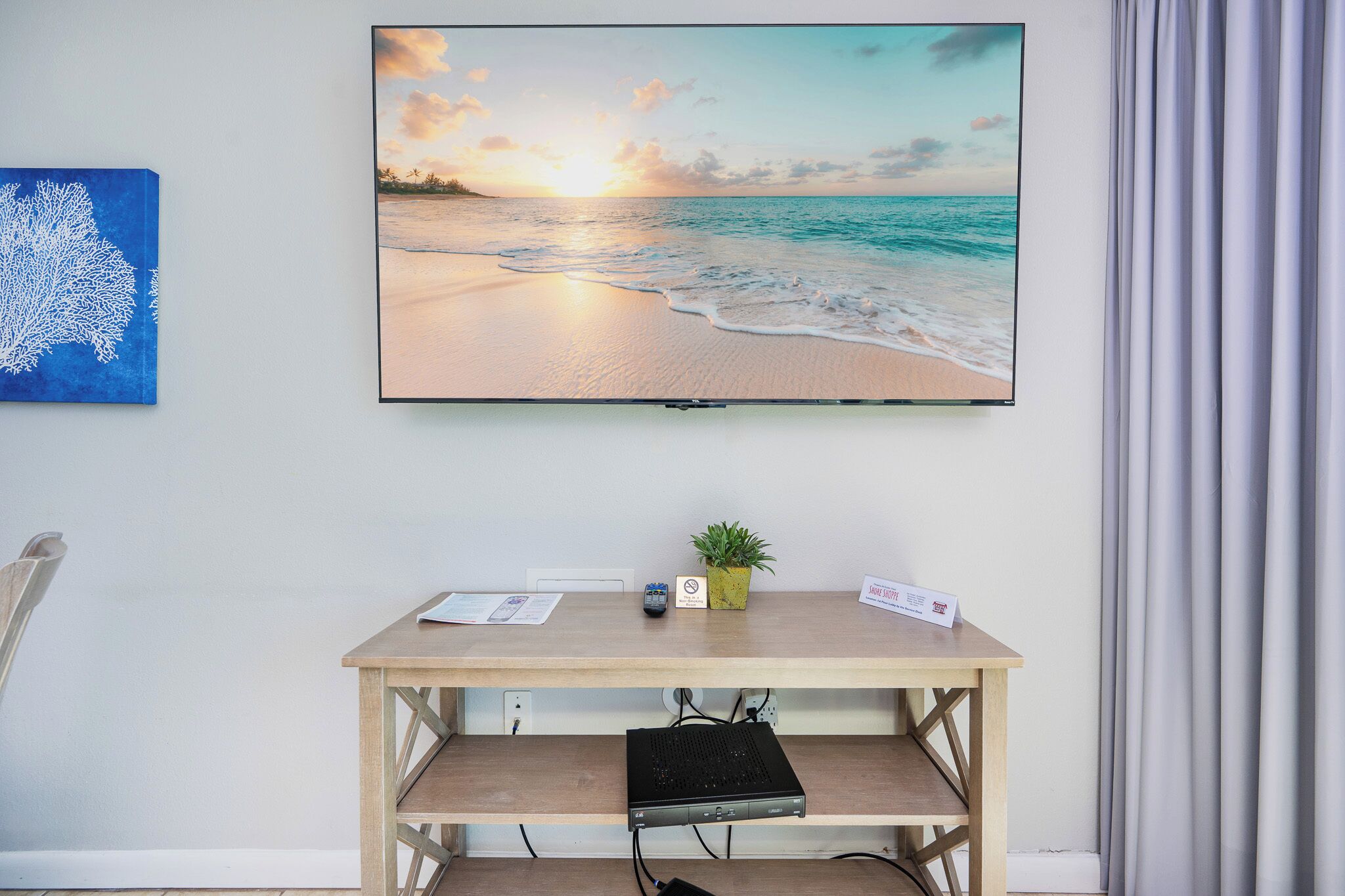 A room with a flat-screen TV displaying a beach scene, a small wooden desk with a plant, and a name card on it.