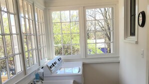 A laundry room with a wooden bench, a washing machine, and a dryer. There are large windows providing natural light and a view of trees. - Haywood House: West Asheville's Newly Restored And Most Walkable Rental! (Asheville)