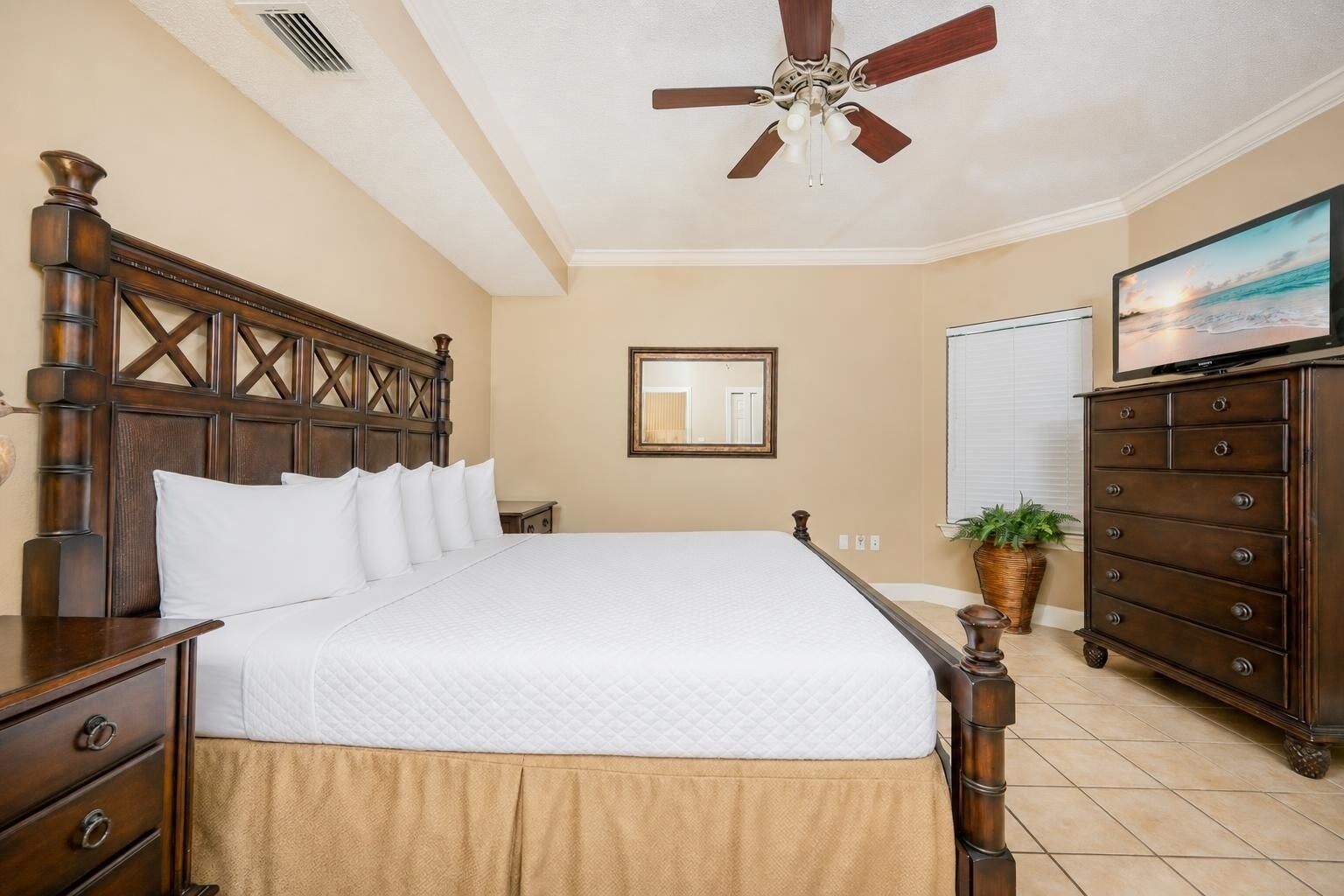 A bedroom with a large bed, wooden headboard, a ceiling fan, a dresser, a television, and a potted plant.