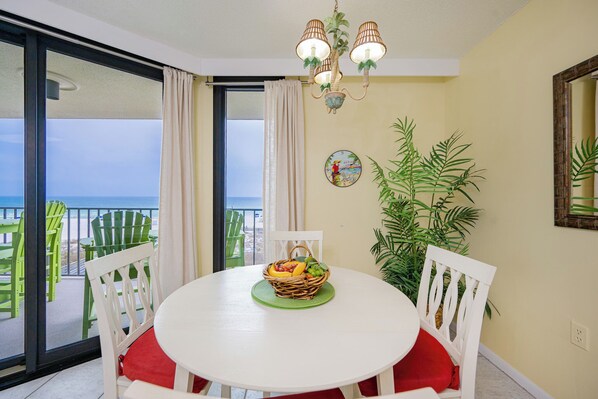 A dining area with a round table, white chairs, a wicker fruit basket, and a view of the ocean through a large window. - Charming 1BR Gulf Front Condo - Phoenix III 3022 (Orange Beach)