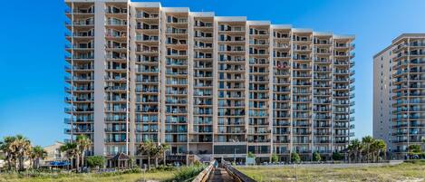 Un hotel de gran altura con varios balcones, un paseo marítimo de madera que conduce a la entrada y un cielo azul despejado.