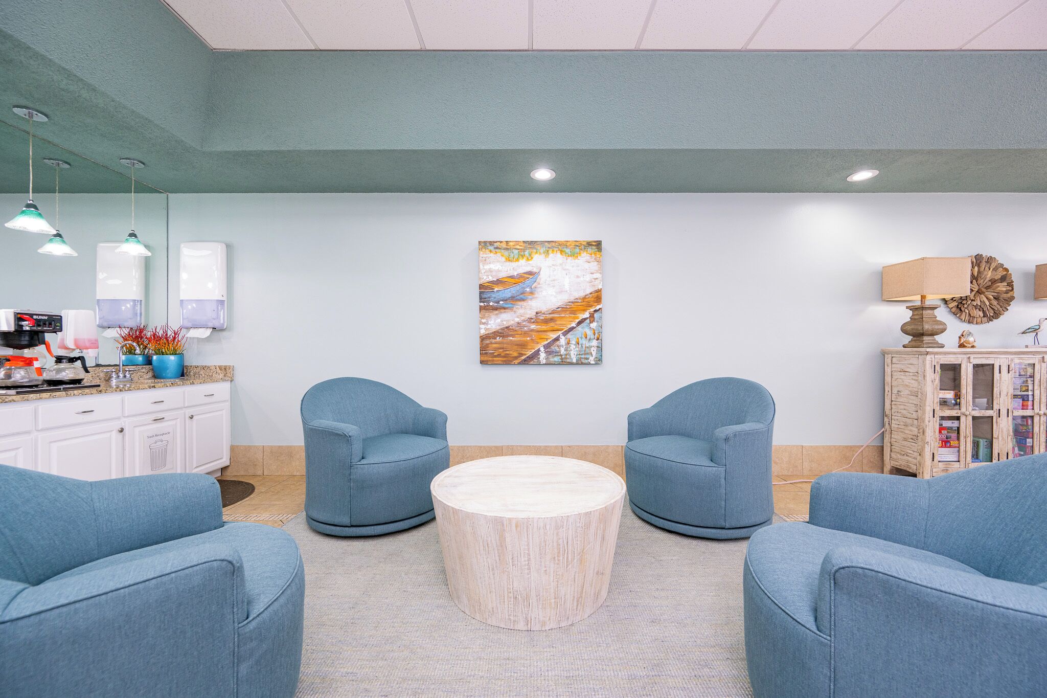 A reception area with a reception desk, two blue armchairs, a round wooden coffee table, a painting on the wall, and a cabinet with books and decorative items.