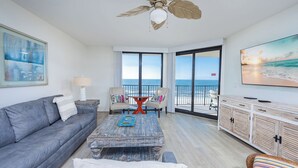 A living room with a gray sofa, a coffee table, and a TV on a wooden cabinet. There is a balcony with a view of the ocean.