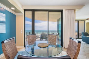 Une table à plateau en verre, des chaises en osier, une vue sur l’océan par une porte-fenêtre coulissante et un balcon avec une chaise bleue.