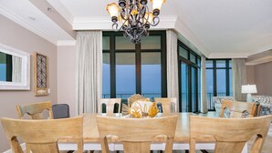 Un coin repas avec une table et des chaises en bois, un lustre et une vue sur l’océan grâce à de grandes fenêtres.