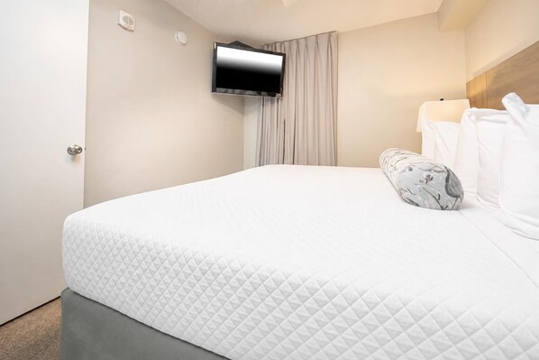 A hotel room with a bed, a flat-screen TV mounted on the wall, a window with curtains, and a bedside table with a lamp.