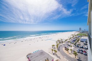 Vista de la playa con un estacionamiento, palmeras y un edificio al fondo.