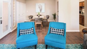 A living room with two blue armchairs, a dining table with chairs, a painting on the wall, and a kitchen area visible through an open door. - Architect-Designed Retreat with a BBQ Patio and Pool (Mesa)